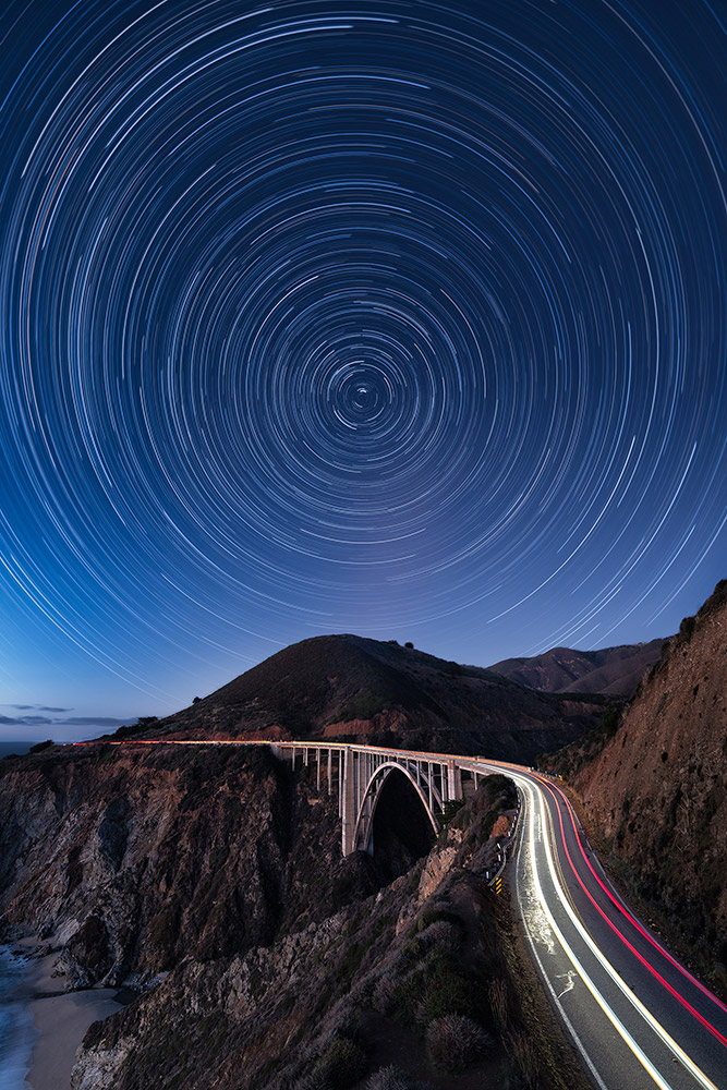 The night sky spinning around the North Star while cars drive through the Bixby Bridge on the Pacific Coast Highway in Central California (USA) by Marcin Zajac